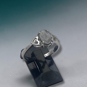 May include: A silver ring with a light gray oval gemstone. The ring features a heart-shaped design on the sides of the band. The ring is displayed on a black stand against a gradient background of teal and white.