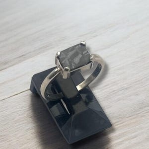 May include: A silver ring with a large, square, gray gemstone. The ring is displayed on a small, black, angled stand, set against a light wood-grain surface. The ring's design is simple, focusing on the prominent stone.