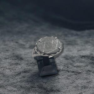 May include: A silver ring with a large, faceted, light-colored gemstone. The ring is set on a dark, geometric base, and the background is a textured, gray surface.