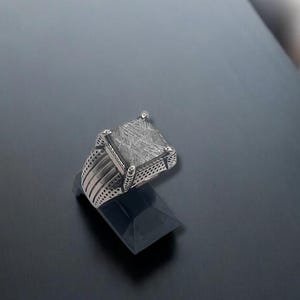 May include: A silver ring with a large, square, textured stone. The ring has a detailed band with multiple lines and a decorative edge around the stone. The ring is displayed on a black stand against a dark background.