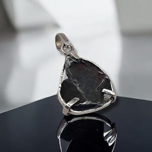 May include: A silver pendant with a dark, irregularly shaped stone. The stone has a textured surface and is held in place by a silver setting. The pendant is displayed on a reflective black surface.