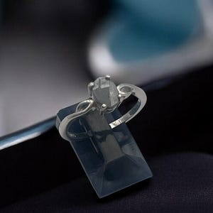 May include: A silver ring with a large, oval-shaped, gray gemstone. The ring is displayed on a dark blue stand inside a black box. The ring's band features a delicate, swirling design. The background is a blurred mix of blue and black.