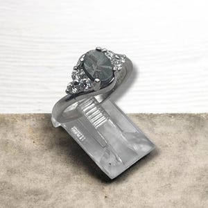 May include: A silver-toned ring featuring a central oval-shaped dark gray stone flanked by three small clear stones on each side. The ring is displayed on a clear plastic stand, showcasing the metallic band and the faceted gemstone arrangement.