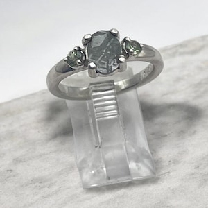 May include: A silver-toned ring featuring a central oval-shaped grey stone flanked by two smaller, round, light green stones. The band is smooth and metallic, holding the three stones in a prong setting, displayed on a clear plastic stand.