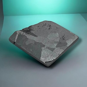 Pode incluir: Uma laje de pedra plana de formato quadrado com uma superfície cinza e carvão manchada, apresentando inclusões cristalinas visíveis e padrões minerais naturais. O objeto possui uma borda texturizada levemente irregular e acabamento fosco.