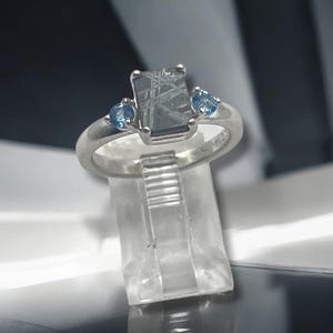 May include: A silver ring featuring a rectangular, grey, needle-like inclusion gemstone at the center, flanked by two small, round, light blue stones. The band is smooth and polished, showcasing a classic three-stone setting design.