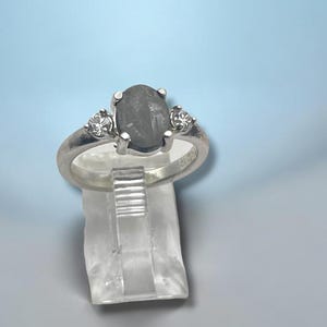May include: A silver ring featuring a central oval-shaped grey stone held by four prongs, flanked by two small round clear stones on each side. The band is polished silver, and the ring is displayed on a clear, vertical acrylic stand.