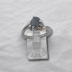 May include: A silver-toned ring featuring a pear-shaped dark gray stone with a visible linear pattern and a smaller round orange stone set beside it. The ring band is smooth and polished, showcasing a bypass design that holds both gemstones securely.