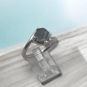 May include: A silver-toned ring featuring a large, dark, oval-shaped center stone held by four prongs. A single, small, clear, round-cut accent stone is set into the band on one side of the main setting, creating an asymmetrical design.