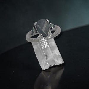 May include: A silver-toned ring featuring a central pear-shaped dark gray stone held by four prongs, flanked by two smaller round dark gray stones on each side. The band is smooth and metallic, showcasing a polished finish throughout the design.