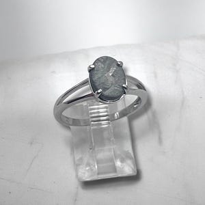 May include: A silver-toned ring featuring an oval-shaped, dark gray stone with a textured, crystalline surface pattern. The stone is held in a four-prong setting atop a split-shank band, creating a clean and polished appearance for the jewelry piece.