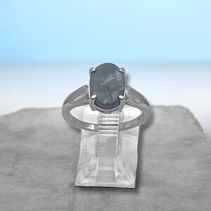 May include: A silver-toned ring featuring a central oval-shaped dark gray stone with visible internal inclusions. The metal band has a split-shank design that connects to a four-prong setting holding the stone securely in place.