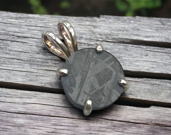 May include: A round, silver pendant with a dark gray meteorite stone set in a silver bezel. The pendant has a silver bail with a double loop.