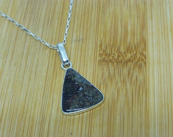 May include: A silver necklace with a triangular pendant. The pendant has a dark, textured surface with a silver border. The chain is a delicate silver link style, resting on a light brown wooden surface.
