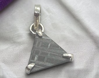 May include: A silver pendant featuring a triangular piece of meteorite with a unique pattern. The pendant has a silver bail for attaching to a chain.