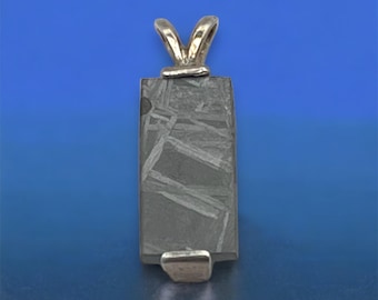 May include: A silver pendant featuring a rectangular piece of meteorite with a Widmanstätten pattern. The pendant is on a blue background.