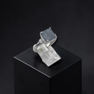 May include: A silver ring featuring a square-cut dark gray meteorite stone with a distinct Widmanstätten pattern. The band is accented with a small, clear, round-cut gemstone on one side, held in a four-prong setting atop a polished metal shank.
