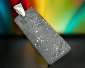 May include: A silver pendant featuring a rectangular piece of meteorite with a unique pattern of lines and swirls. The pendant is made of a dark gray metal and has a silver bail.