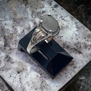 Puede incluir: Un anillo de plata con una piedra gris ovalada engastada en una banda de plata decorativa.