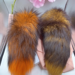 Può includere: Due code di volpe soffici, una arancione e marrone, l'altra marrone e dorata. Le code sono esposte su una superficie rosa chiaro.