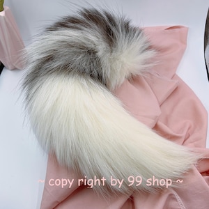 Può includere: Una coda di pelliccia sintetica bianca e grigia, soffice, con una lunga punta bianca e fluida. La coda è appoggiata su una superficie di tessuto rosa.