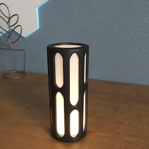 K&ouml;nnte beinhalten: Eine schwarze zylindrische Tischlampe mit einer wei&szlig;en Lichtquelle und einem geometrischen Ausschnittmuster.