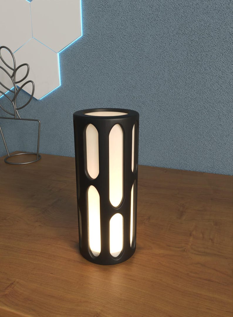 K&ouml;nnte beinhalten: Eine schwarze zylindrische Tischlampe mit einer wei&szlig;en Lichtquelle und einem geometrischen Ausschnittmuster.