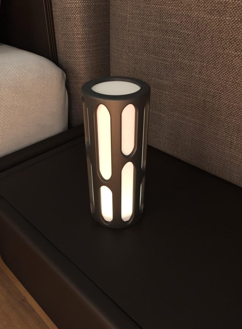 K&ouml;nnte beinhalten: Eine schwarze zylindrische Lampe mit einer wei&szlig;en Lichtquelle und einem geometrischen Design. Die Lampe steht auf einer dunkelbraunen Oberfl&auml;che.