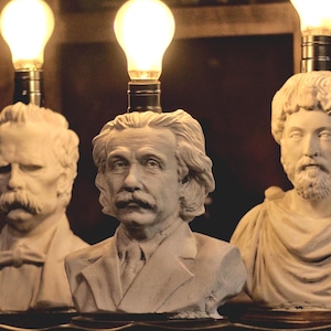 May include: A collection of bust lamps featuring famous historical figures. The lamps have a light bulb on top of the bust. The busts are made of a light-colored material and are set against a dark background.
