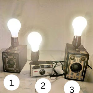Könnte beinhalten: Drei Vintage-Kameras, die zu Lampen umfunktioniert wurden, jede mit einer leuchtenden weißen Glühbirne an der Oberseite. Die Kameras sind schwarz und silber, wobei das mittlere Gerät als Kodak Instamatic 104 und das rechte als Brownie Junior Six-16 beschriftet ist.