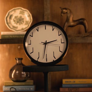 Könnte beinhalten: Eine runde Uhr mit schwarzem Rahmen und weißem Zifferblatt, auf einem schwarzen Ständer. Der Hintergrund zeigt ein Holzregal mit einem dekorativen Teller, Büchern und einer Pferdefigur. Der Durchmesser der Uhr beträgt ca. 25 cm.