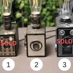 Può includere: Tre fotocamere a cassetta vintage riutilizzate come lampade da tavolo, ciascuna sormontata da una lampadina in stile Edison. La fotocamera a sinistra è una Brownie Junior, quella centrale una Ansco Shur-Flash e quella a destra una Brownie Target Six-16. Le etichette Sold appaiono sulle fotocamere 1 e 3.