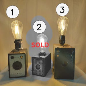 Puede incluir: Tres lámparas de cámara vintage con bombillas estilo Edison. Las lámparas están hechas de cámaras antiguas en tonos negros y grises. Una lámpara tiene el texto "SHUR SHOT" en la parte delantera. La lámpara del medio está marcada como "SOLD".