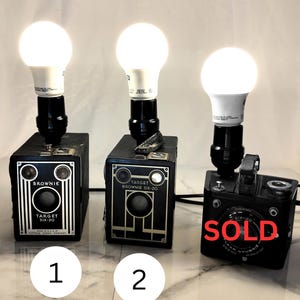 Puede incluir: Tres cámaras de caja negras vintage reutilizadas como lámparas de mesa, cada una coronada con una bombilla LED blanca brillante. Las cámaras están etiquetadas como Brownie Target Six-20 y Six-20 Flash. La tercera cámara a la derecha tiene la palabra SOLD en rojo.