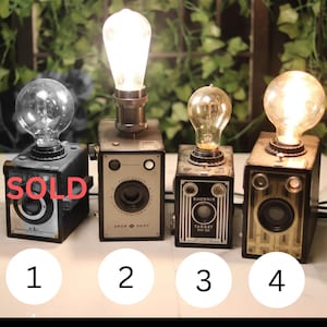 Puede incluir: Cuatro cámaras de caja antiguas reconvertidas en lámparas de mesa, cada una coronada con una bombilla brillante estilo Edison. Las cámaras están numeradas del 1 al 4, con la primera marcada con el texto rojo SOLD. Cada unidad tiene un cuerpo único.