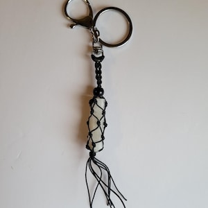 May include: A handmade keychain featuring a light-colored stone encased in a black macrame net. The keychain has a silver-colored clasp and ring, with a black beaded accent and a tassel of black cords at the bottom.