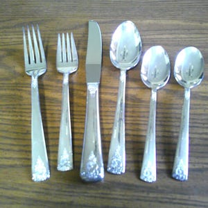 May include: A set of six silver-toned stainless steel flatware pieces arranged in a row, including a dinner fork, a salad fork, a dinner knife, a large soup spoon, and two smaller teaspoons, all featuring a decorative floral pattern on the handles.