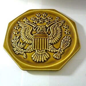 May include: A gold-colored, octagonal plate with a detailed embossed design. The plate features a central eagle emblem with outstretched wings, surrounded by stars and the words "E PLURIBUS UNUM". The plate has a slightly raised edge.