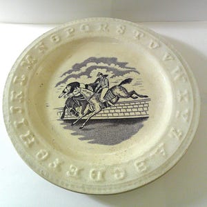 May include: Cream-colored ceramic plate with a black and white illustration of a horse race. The plate's rim features embossed letters of the alphabet. The illustration depicts two horses jumping over a brick wall, with riders.