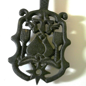May include: A dark, cast-iron trivet with a handle and decorative cut-out design. The trivet features a central shape with a star, two stylized brush-like shapes, and other ornate elements. The handle has a textured pattern.
