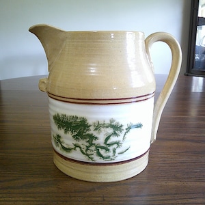 May include: A tan ceramic pitcher with a curved handle and spout. The pitcher features a white band with a green pine tree design, and brown stripes. The pitcher is likely used for serving beverages.