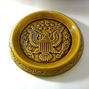May include: A round, ceramic dish in a golden-yellow hue. The dish features a detailed embossed design of the American Great Seal, including an eagle, stars, and the words "E Pluribus Unum". The rim is decorated with a floral pattern.