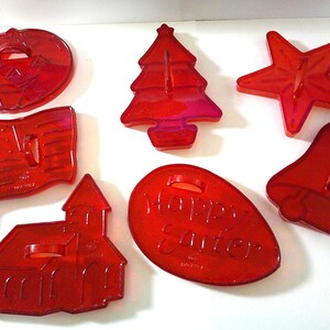 May include: A collection of red plastic cookie cutters in various shapes, including a pumpkin, Christmas tree, star, American flag, church, Easter egg with "Happy Easter" text, and a bell. These cutters are designed for festive baking.