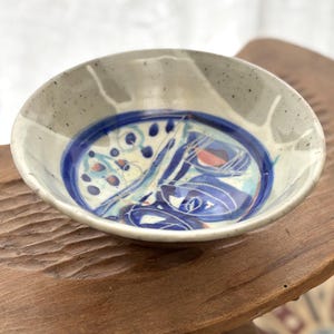 Peut inclure: Un petit bol en céramique fait à la main avec un extérieur gris moucheté et un design intérieur bleu et blanc vibrant. Le bol présente un bord bleu foncé et des motifs abstraits dans les tons de bleu, de rouge et de blanc. Il repose sur une surface en bois.