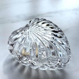 May include: A clear, heart-shaped glass trinket box with a ridged, fan-like design. The lid and base have a scalloped edge. The box is sitting on a white surface, reflecting light and creating shadows.