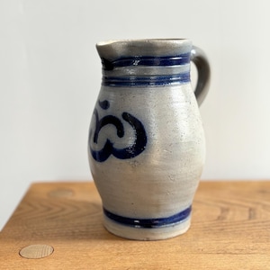 Peut inclure: Une cruche en grès aux tons gris et bleus. La cruche est dotée d'une anse et est ornée de bandes bleu foncé et d'un motif floral stylisé. Elle repose sur une surface en bois.