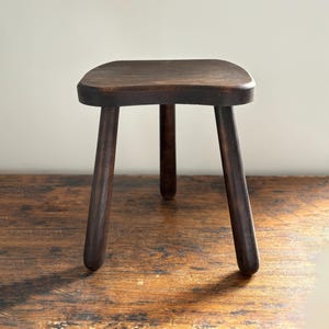 May include: A dark brown, three-legged wooden stool with a square seat and rounded corners. The cylindrical legs taper towards the floor. The stool is set on a wooden surface.