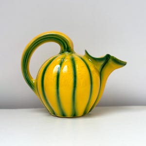May include: A ceramic pitcher with a yellow body and green vertical stripes. The handle and spout are also green. The pitcher has a rounded shape and sits on a white surface. The pitcher is likely used for serving beverages.