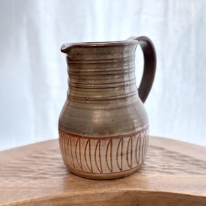 Puede incluir: Jarra de cerámica hecha a mano con asa y boquilla marrón oscuro. La jarra tiene crestas horizontales y un patrón decorativo de líneas verticales en un color marrón claro cerca de la base. La jarra es de color neutro, con una apariencia rústica y artesanal.