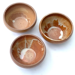 May include: Three small, handmade ceramic bowls. The bowls are a warm brown color with a speckled texture and a lighter, off-white interior. The bowls have a rounded shape and are arranged on a white surface.